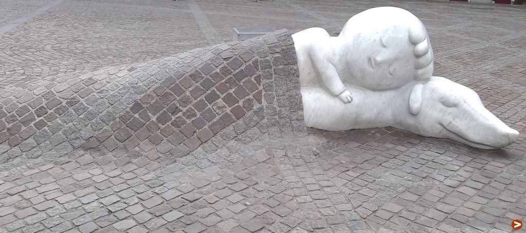 White stone statue of a small boy lying down and hugging his dog. The cobblestone paving has been fashioned to look like a blanket