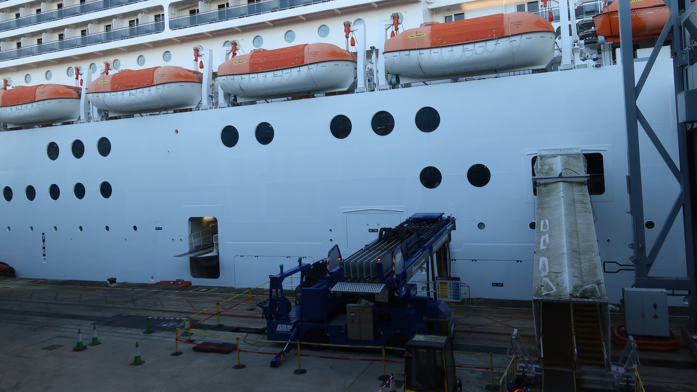 A large piece of equipment sits on the dockside, a wide arm extending to the white ship behind and carrying several big cables