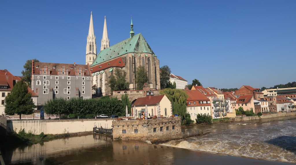 A large Gothic church rises above old buildings, a wide river rushes over a weir in the foreground